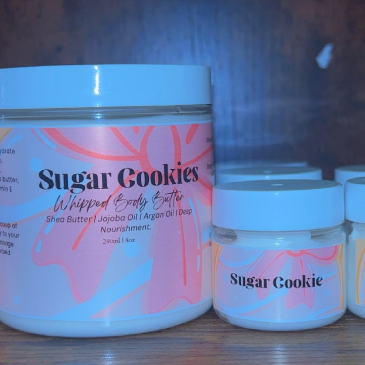 Jar of 'Sugar Cookies' whipped body butter with smaller jars in the background on a wooden surface.